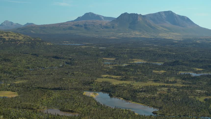 A wide, static aerial shot of a vast, untouched wilderness valley with lush forests and rivers below a distant mountain range