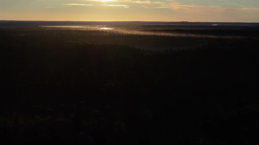 The sun sets over a vast, silhouetted forest landscape, casting a golden glow on a misty river in the distant valley