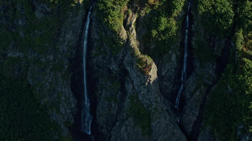 Twin waterfalls carve paths down a steep, verdant cliff face in a serene mountain landscape
