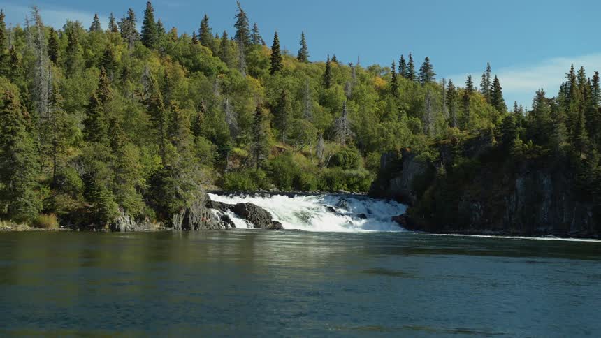 A serene waterfall cascades over rocky tiers into a wide river, nestled within a dense, verdant forest under a clear blue sky