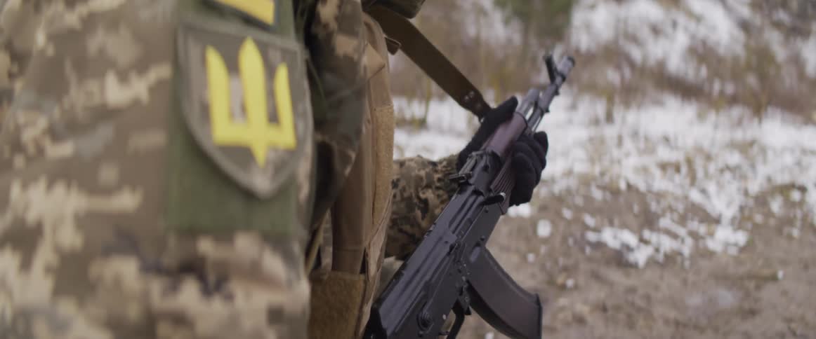 An over-the-shoulder slow-motion shot reveals a Ukrainian soldier holding a Kalashnikov rifle in a wintry setting