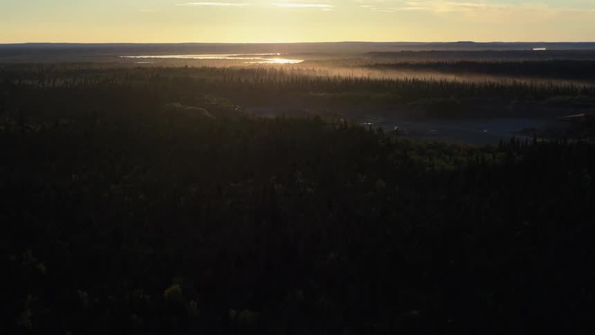 An aerial view slowly pushes in over a vast, dark forest, revealing a misty valley and a gravel pit illuminated by the golden light of sunrise
