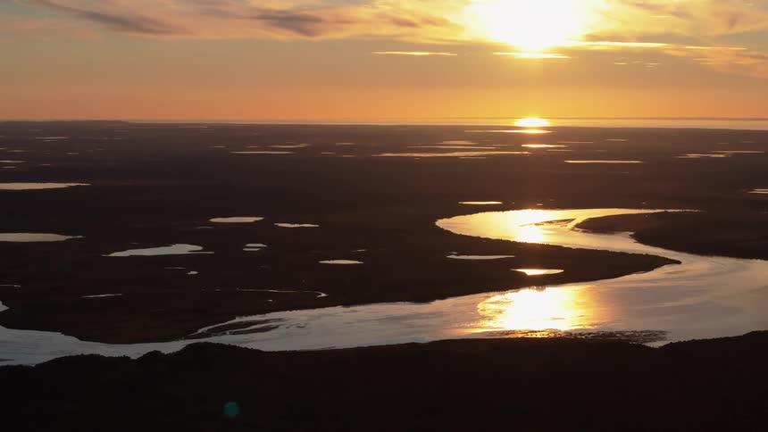 A stunning aerial view shows a vast, watery landscape with a meandering river reflecting the golden light of the setting sun on the horizon