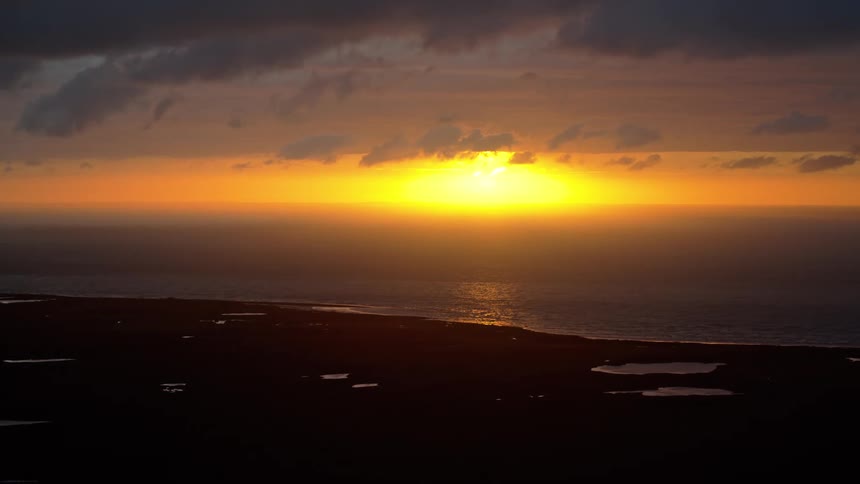 A dramatic, golden yellow sun sets over the ocean horizon, casting the dark coastline and its small lakes into silhouette