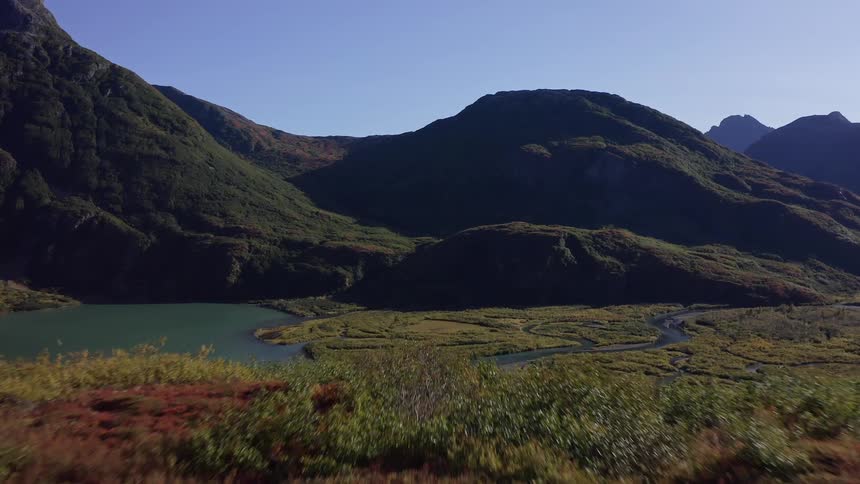 A stunning aerial shot flies over an autumnal hillside to reveal a vast mountain valley with a serene lake and winding river delta under a clear blue sky