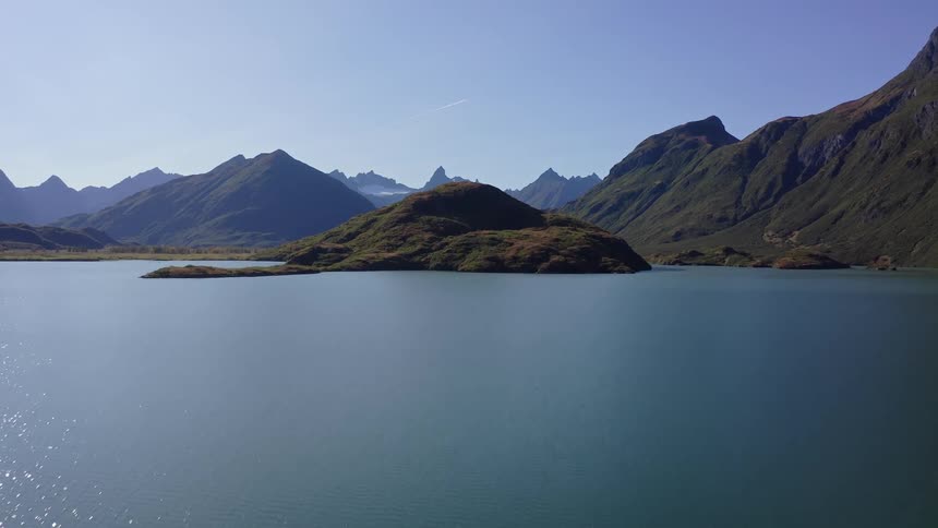 A slow aerial pullback reveals a tranquil alpine lake with a small island, framed by majestic mountains under a clear blue sky