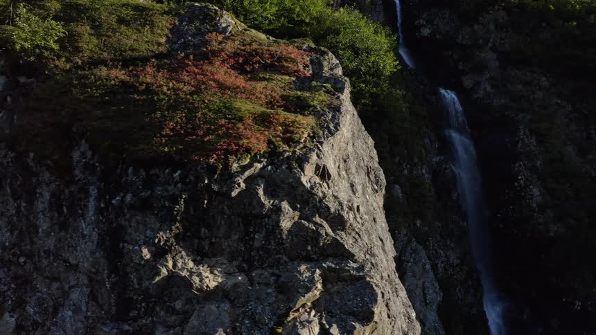 A sunlit rocky cliff with green and red foliage stands next to a cascading waterfall in the mountains