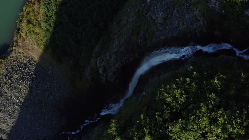 A top-down aerial view slowly reveals a powerful river and waterfall carving through a dense green forest and a dark, rocky canyon