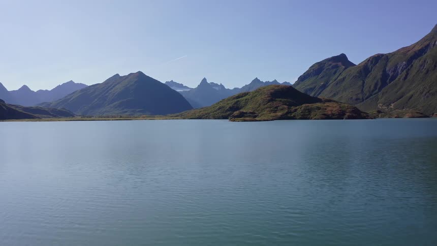 A slow aerial pullback reveals the stunning beauty of a tranquil turquoise lake nestled within a rugged mountain range on a clear day