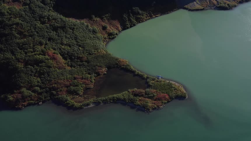 A slow aerial zoom-in reveals a helicopter landed on a scenic peninsula surrounded by a tranquil, green alpine lake