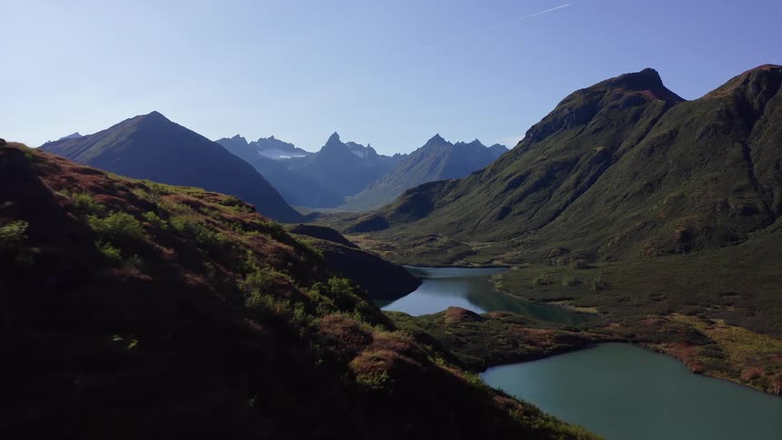An epic aerial shot flies over a colorful, grassy ridge to reveal a breathtaking mountain valley with a tranquil lake on a clear day