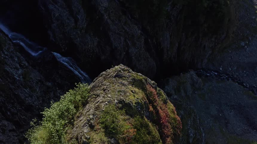Aerial drone shot slowly flies over a sun-kissed, mossy mountain ridge, revealing a deep, shadowy canyon with a cascading waterfall below