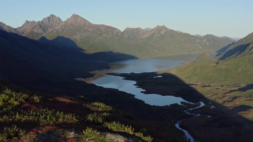 A majestic aerial view shows a serene lake nestled in a vast mountain valley as long shadows stretch across the landscape at sunrise