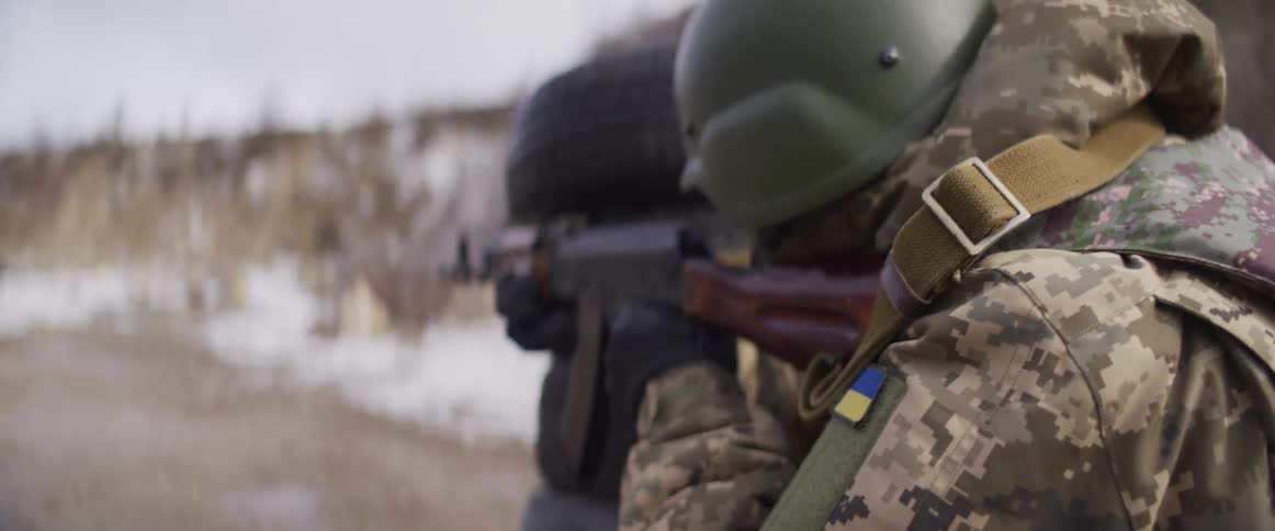 A Ukrainian soldier in winter camouflage aims an assault rifle as the camera slowly pulls focus to the Ukrainian flag patch on his arm