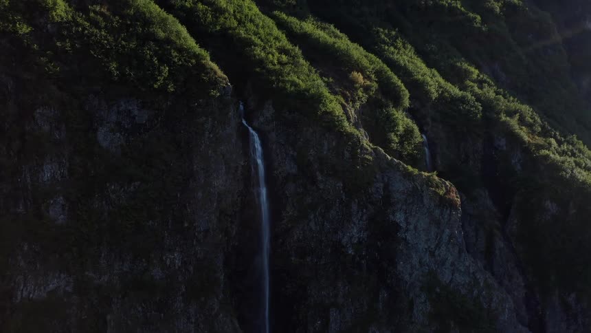 An aerial view rises up a sun-kissed, tree-covered mountain revealing multiple waterfalls cascading down the dark, rocky cliffs