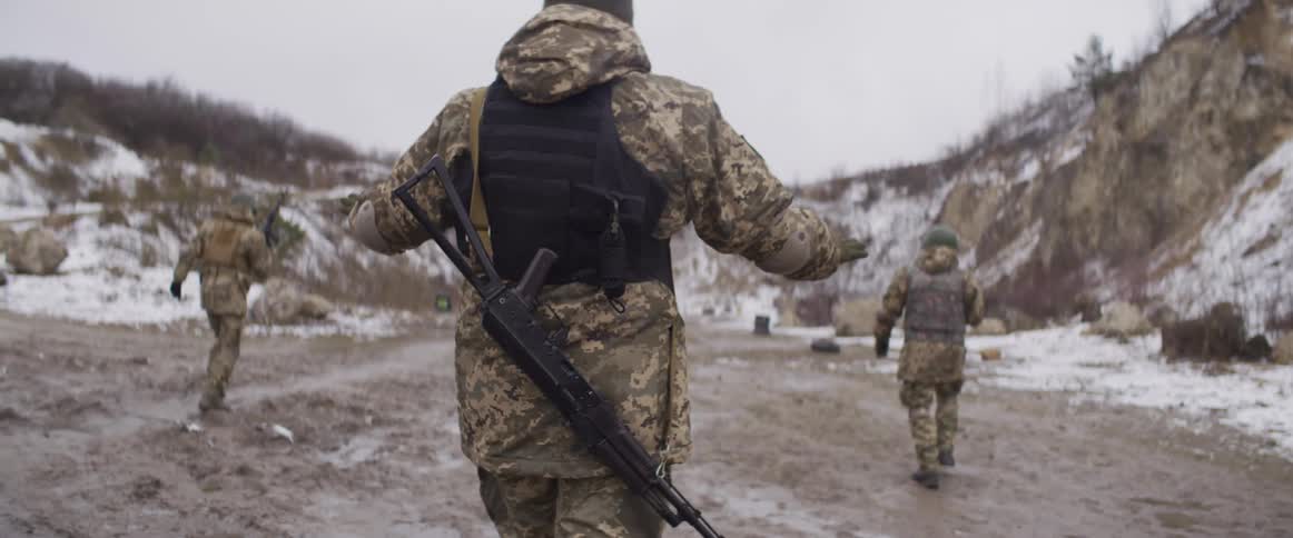 Soldiers in full combat gear with rifles walk through a muddy, snowy landscape in slow motion