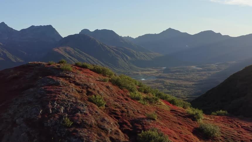 Aerial drone flies over a vibrant red autumn ridge to reveal a stunning mountain valley and lake at sunrise