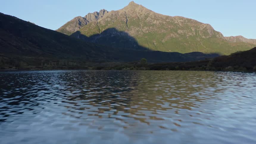 A serene aerial shot slowly pulls back over a calm lake, revealing a majestic mountain range lit by the morning sun