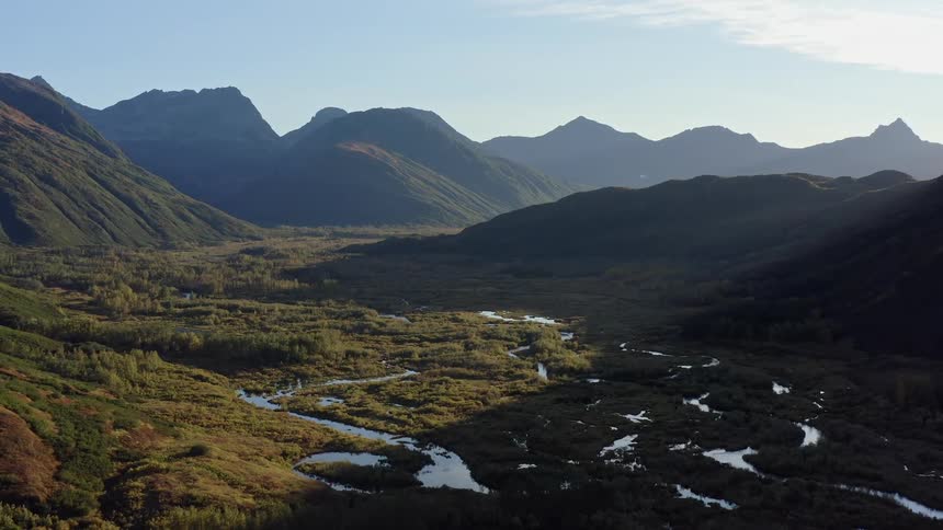 A slow aerial retreat reveals a magnificent mountain valley at sunrise, with a winding river flowing through the wild, green landscape