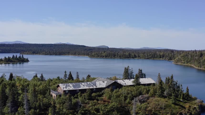 Ascending aerial shot over an old, weathered lodge reveals a vast, tranquil lake surrounded by a dense forest