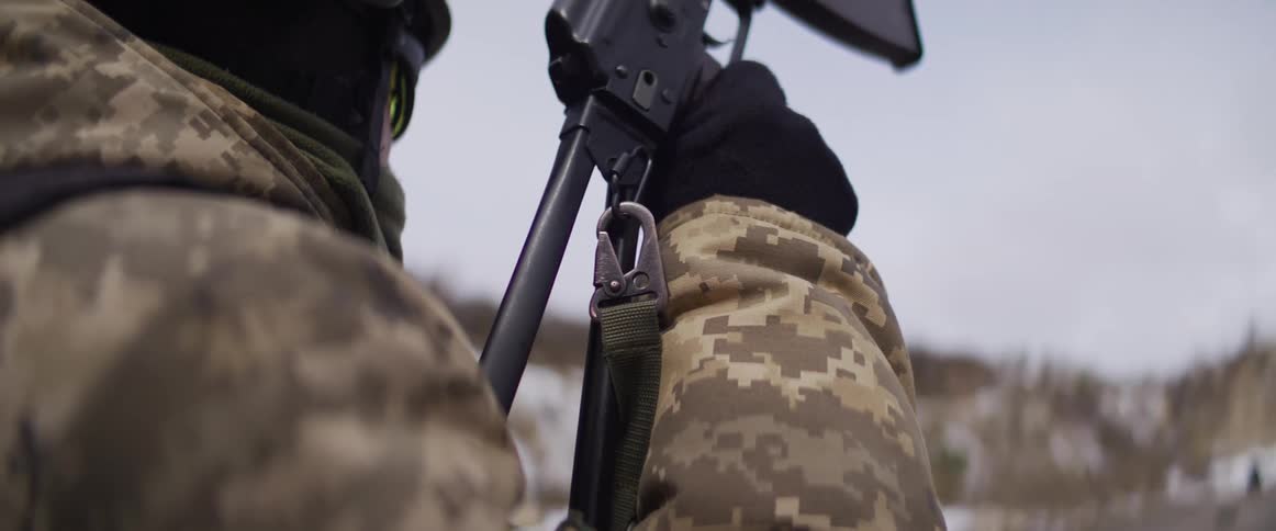 Close-up over-the-shoulder view of a Ukrainian soldier in winter camouflage holding a rifle, on duty in a snowy landscape