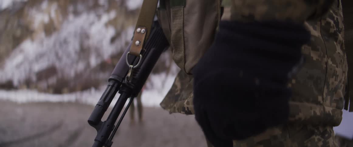 A close-up shot focuses on a soldier in camouflage holding a rifle, standing guard in a cold, snowy landscape as another soldier walks in the background