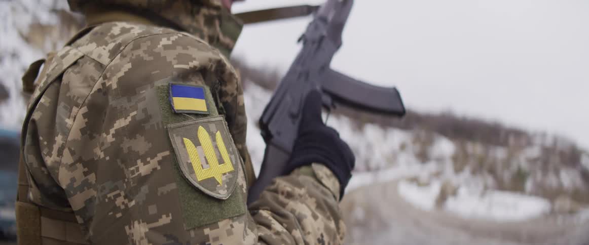 Close-up of a Ukrainian soldier in camouflage holding a rifle with the national flag and coat of arms on their sleeve during winter