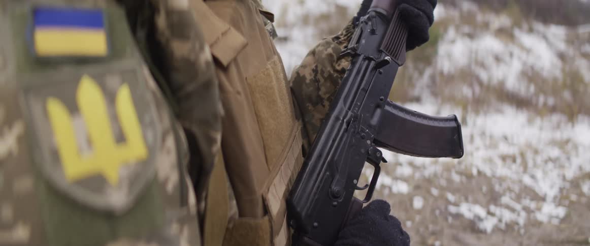 An over-the-shoulder slow-motion shot reveals a Ukrainian soldier in camouflage holding a rifle in a snowy winter landscape