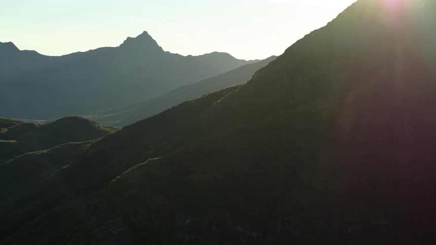 A slow aerial reveal of a vast mountain landscape as the sun sets behind the peaks, casting the green hills into shadow