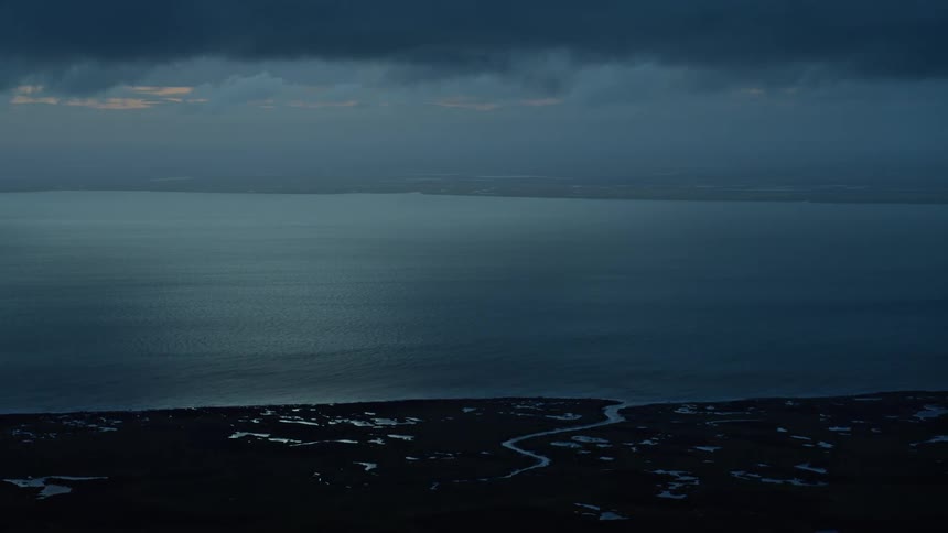 A serene yet moody view of a vast body of water meeting a silhouetted coastline under a dark, cloudy sky