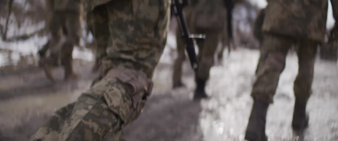 Slow-motion low-angle view of armed soldiers in combat gear running through a wet, muddy battlefield