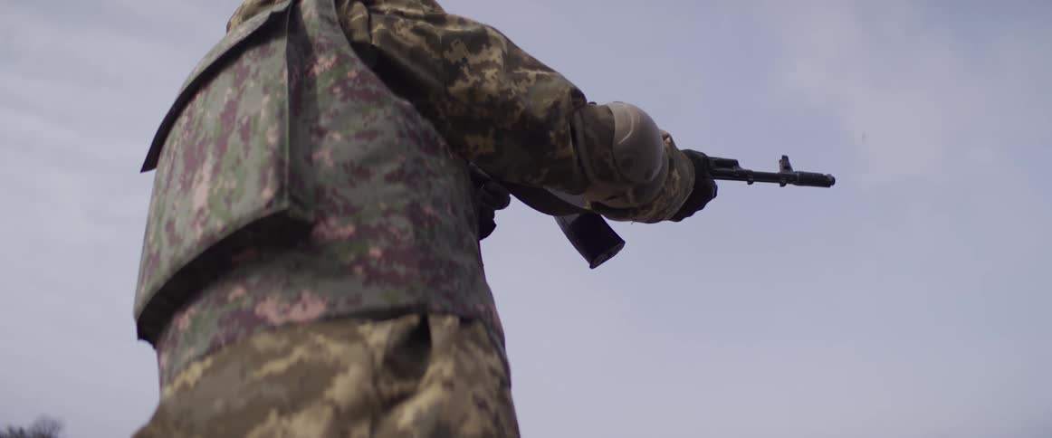 Slow motion footage from behind a soldier in camouflage as he fires an assault rifle, with bullet casings flying out
