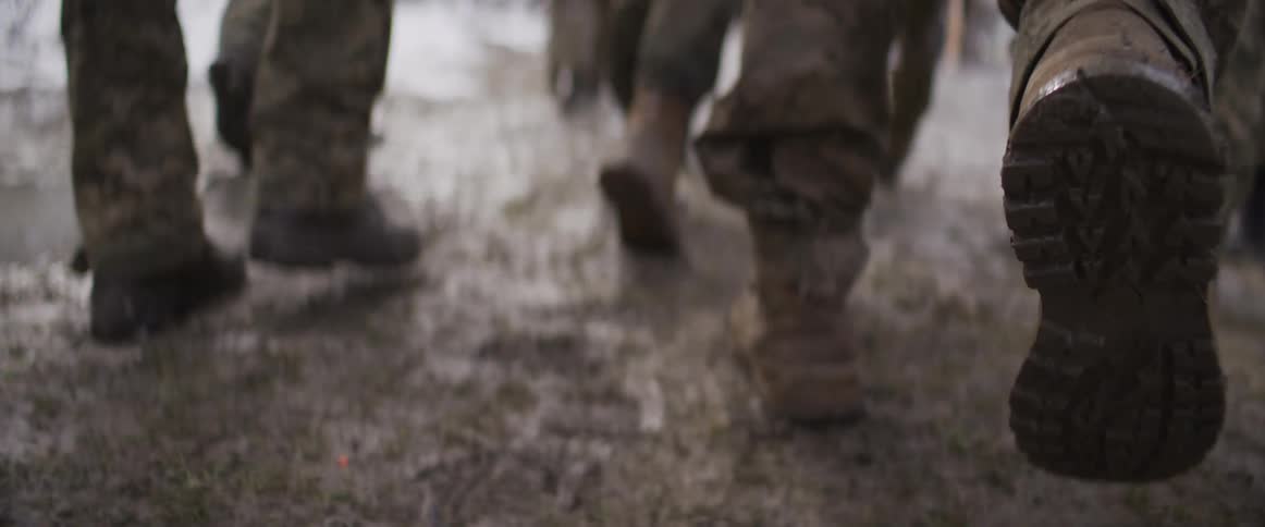 A slow-motion, low-angle shot shows soldiers in camouflage marching through muddy, snowy terrain