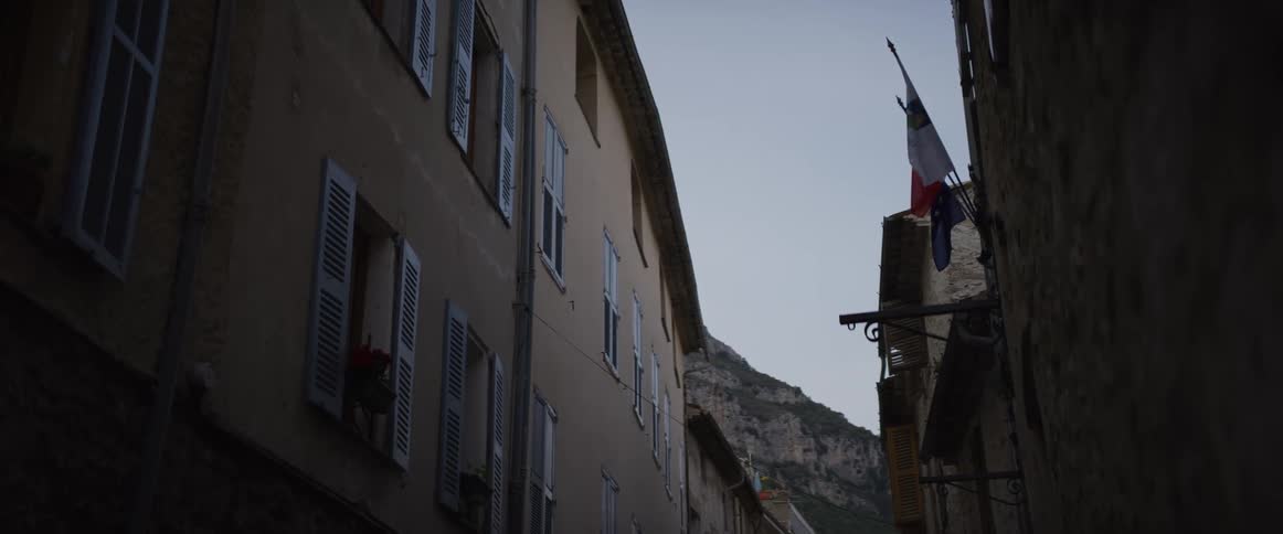 A French flag hangs over a narrow street in an old European village at dusk with mountains in the background