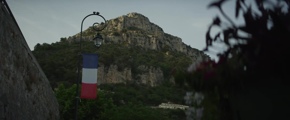 A French flag hangs from a classic lamppost with a beautiful, rugged mountain landscape in the background during golden hour