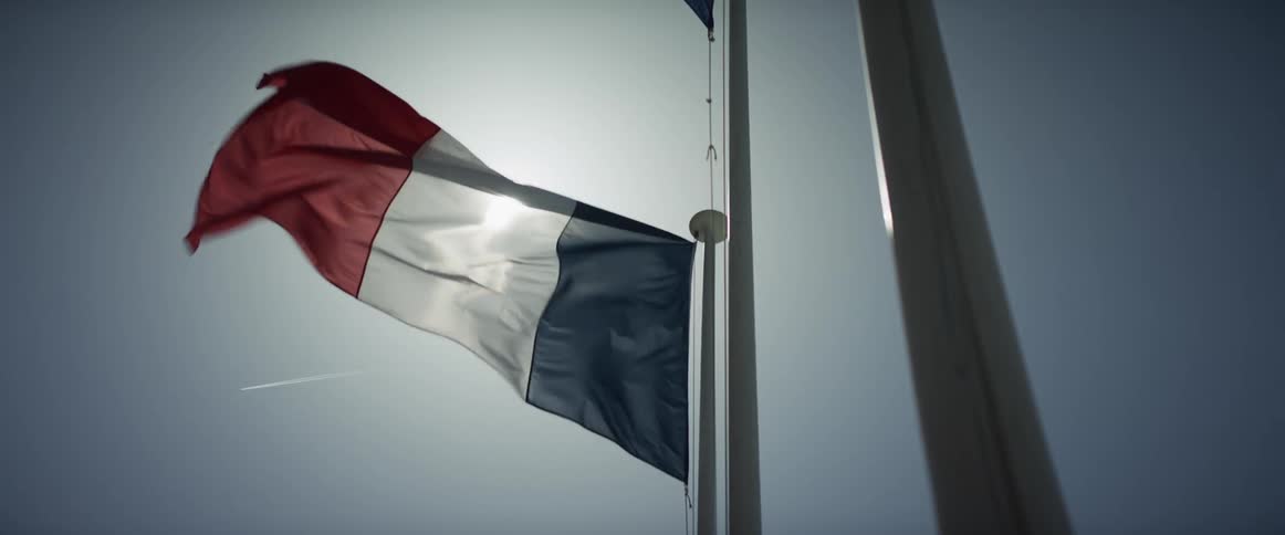 The French flag waves in slow motion at half-mast, backlit by the sun against a hazy sky