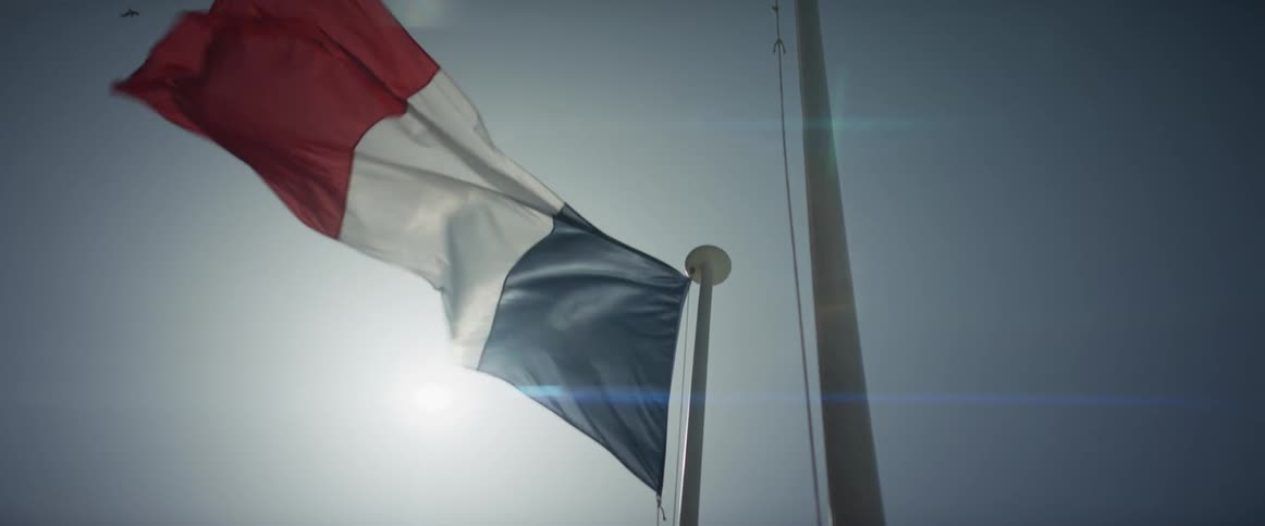 The French flag waves slowly at half-mast, backlit by the sun with a cinematic lens flare