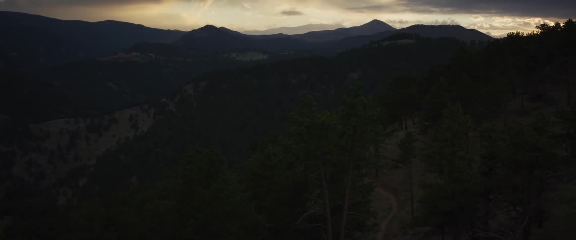 A scenic, moody view of a dark mountain range covered in evergreen trees as the sun sets behind dramatic clouds