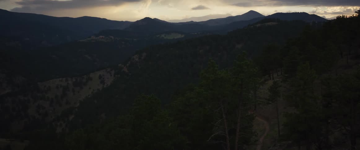 A vast mountain landscape covered in dark forests under a cloudy, dramatic sky at twilight