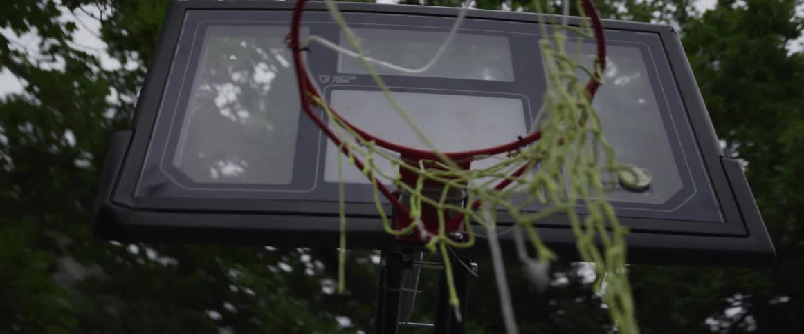 A slow motion orbiting shot of a broken basketball hoop with a tattered net in a backyard on a cloudy day