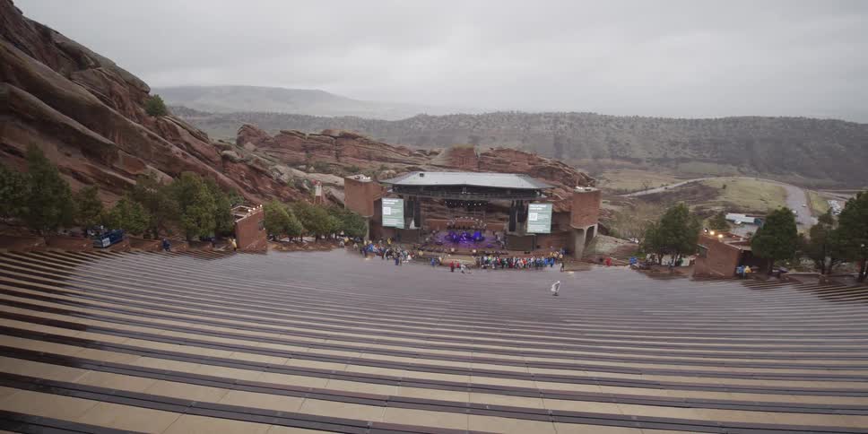 A wide pan of the audience gathering at the stage of the Red Rocks Amphitheater for a concert on a rainy day