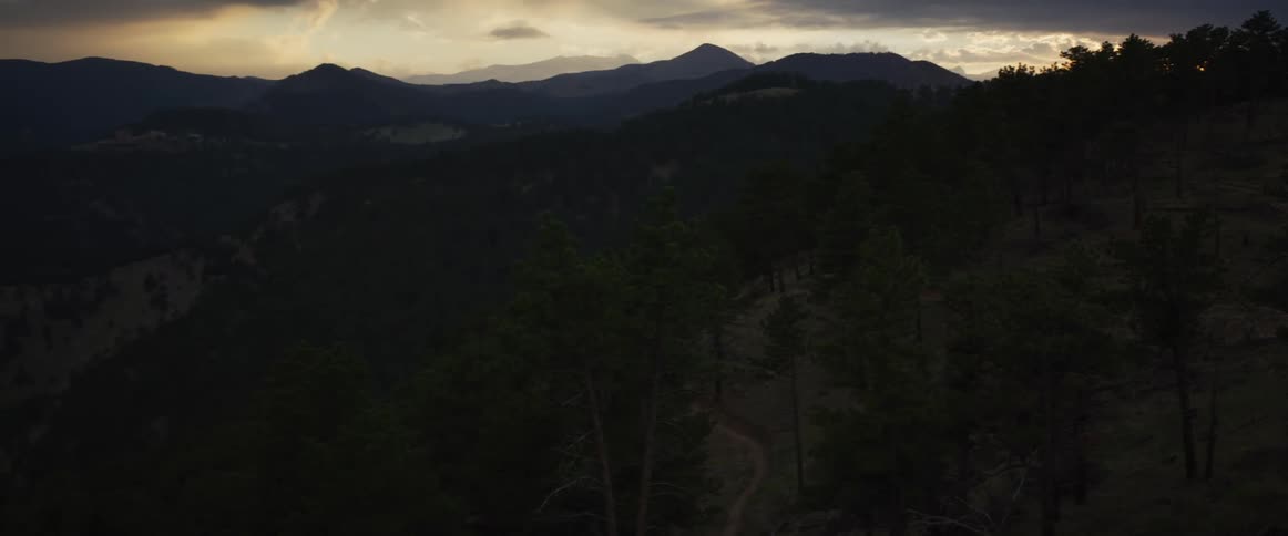 A moody, dramatic view of a dense pine forest and distant mountain range silhouetted against a twilight sky