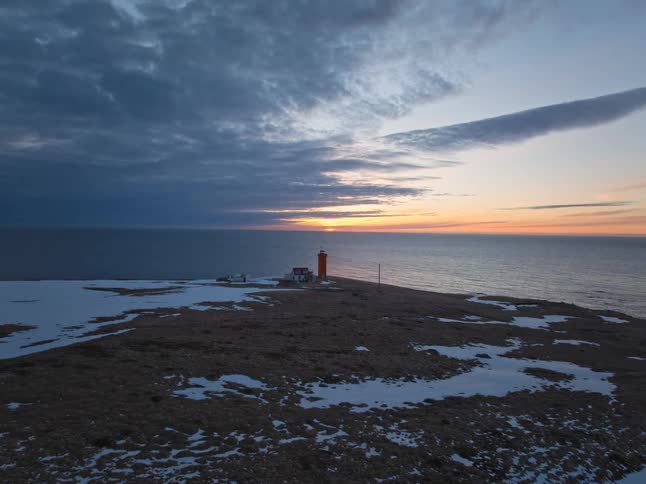 An aerial view pulls back from a vibrant red lighthouse on a snowy, remote peninsula as the sun sets over the calm ocean