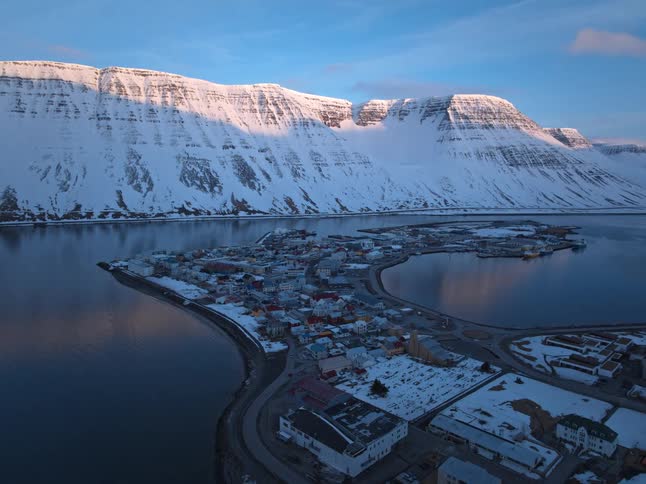 A stunning aerial view of a picturesque coastal town nestled at the base of vast, snow-covered mountains at sunrise