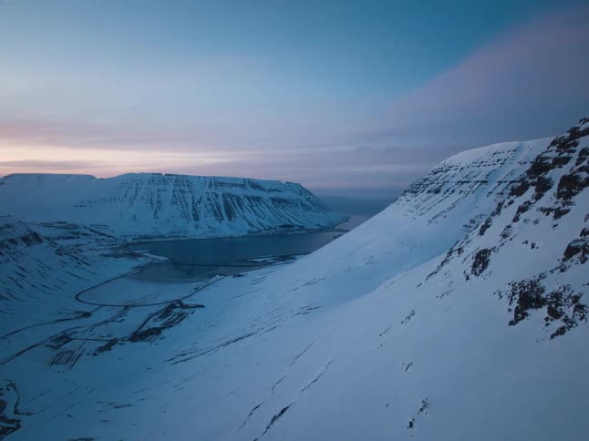 A vast, snowy Icelandic mountain valley and fjord are revealed in a serene aerial shot at dusk