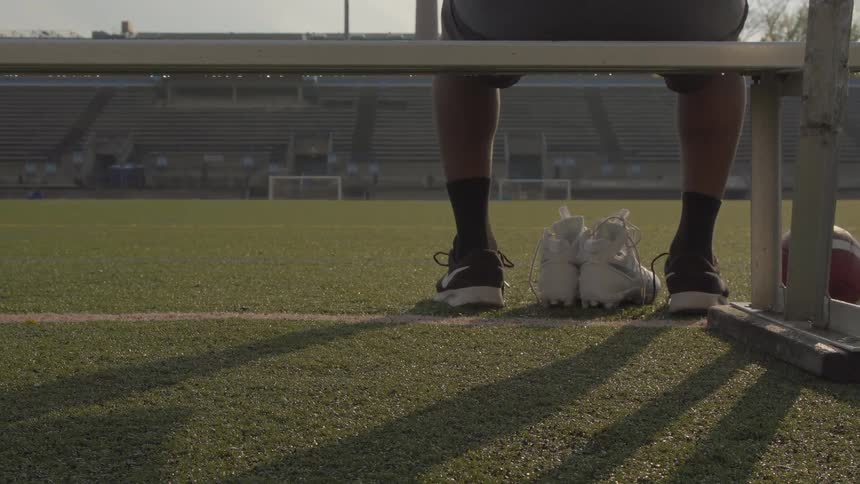 A man sits on a bench by a field with sneakers by his feet