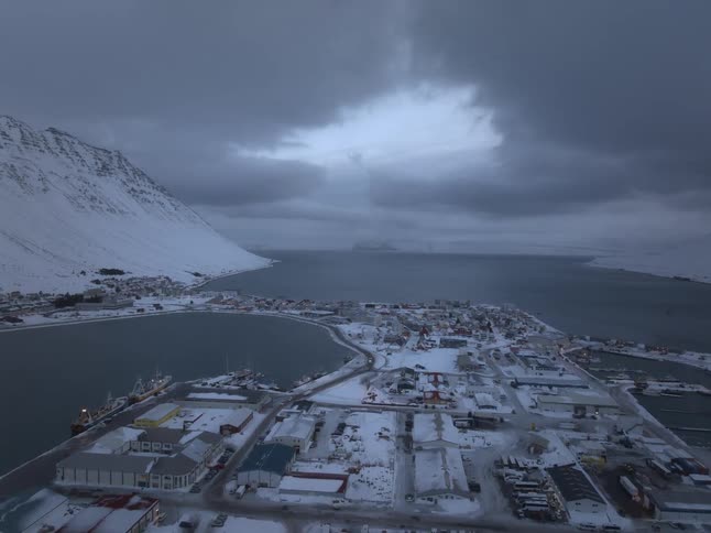 A dramatic aerial view shows a snow-covered coastal town in a fjord under a moody, overcast sky