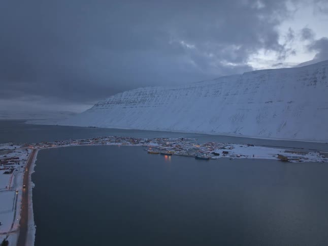 An epic aerial shot pushes towards a remote, snow-covered Icelandic fishing village nestled in a fjord at dusk