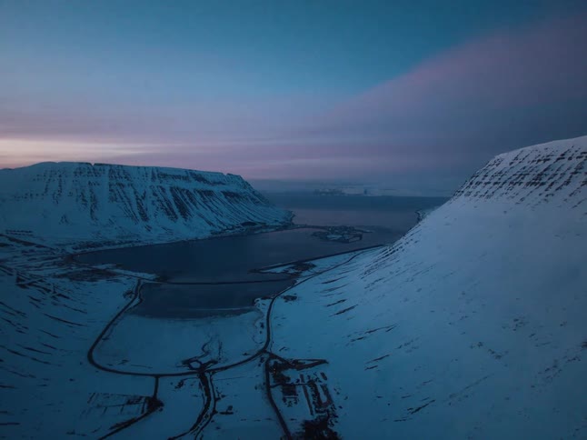 A serene aerial timelapse captures the transition from a colorful sunset to deep twilight over a snow-covered fjord nestled between mountains