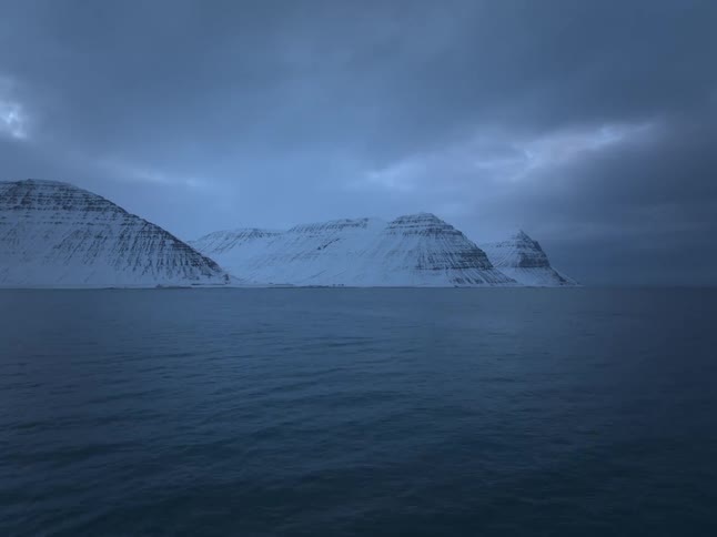 A dramatic wide shot of majestic snow-covered mountains rising from a dark blue ocean under a moody, cloudy sky